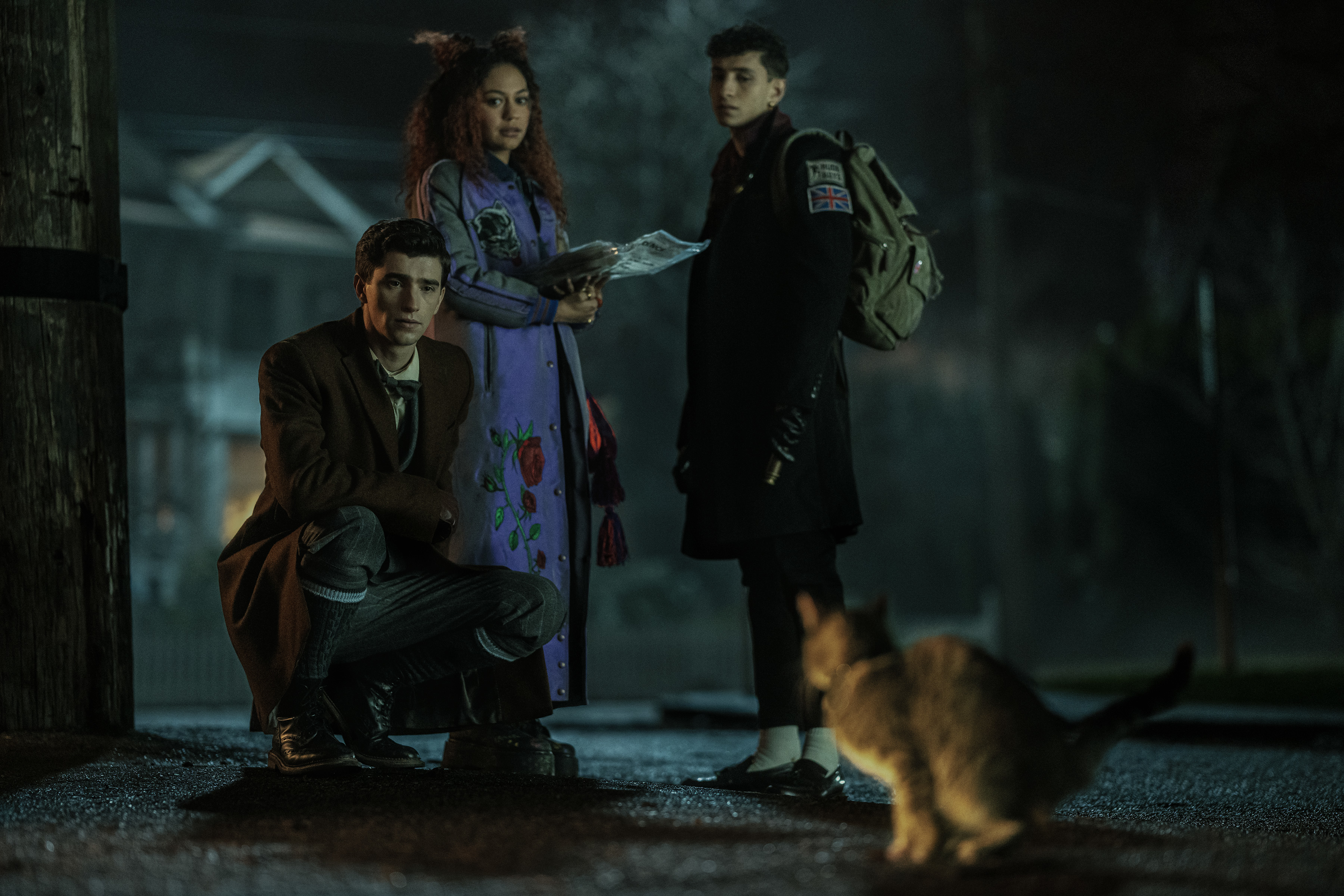 Three individuals, likely actors, in a dimly lit outdoor scene, with one crouching, two standing, and a cat in the foreground. They appear curious or tense