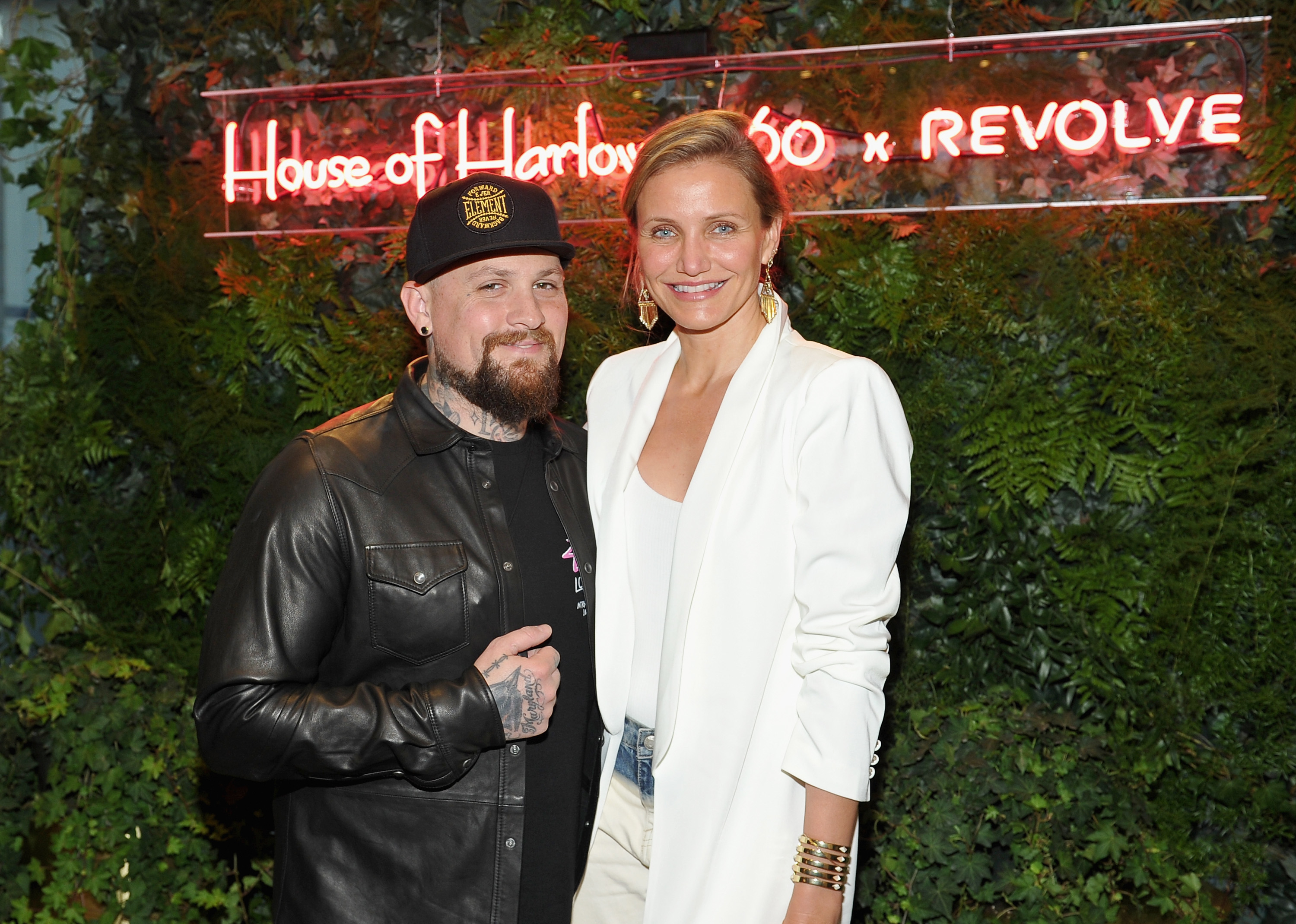 Man in leather jacket and hat with a woman in white blazer at a House of Harlow event
