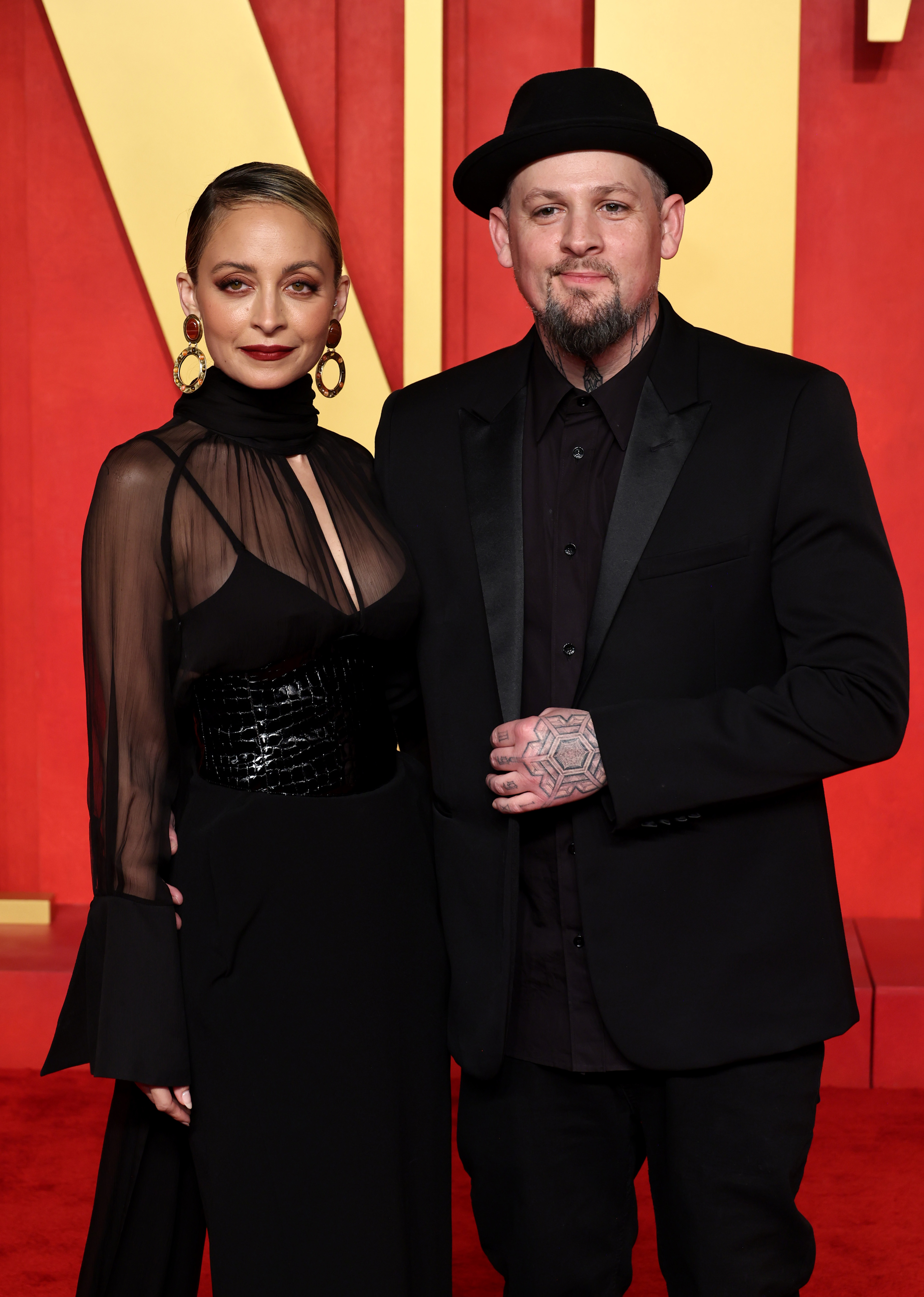 Two people posing, one in a sheer black dress and the other in a suit with a hat, at a formal event