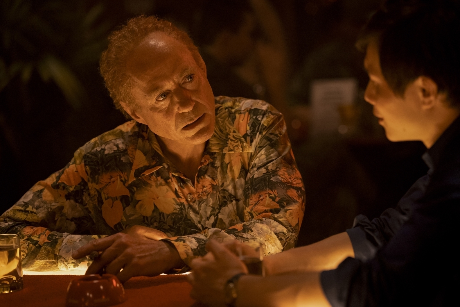 Two characters in a tense scene at a table, one in a floral shirt, from a TV show
