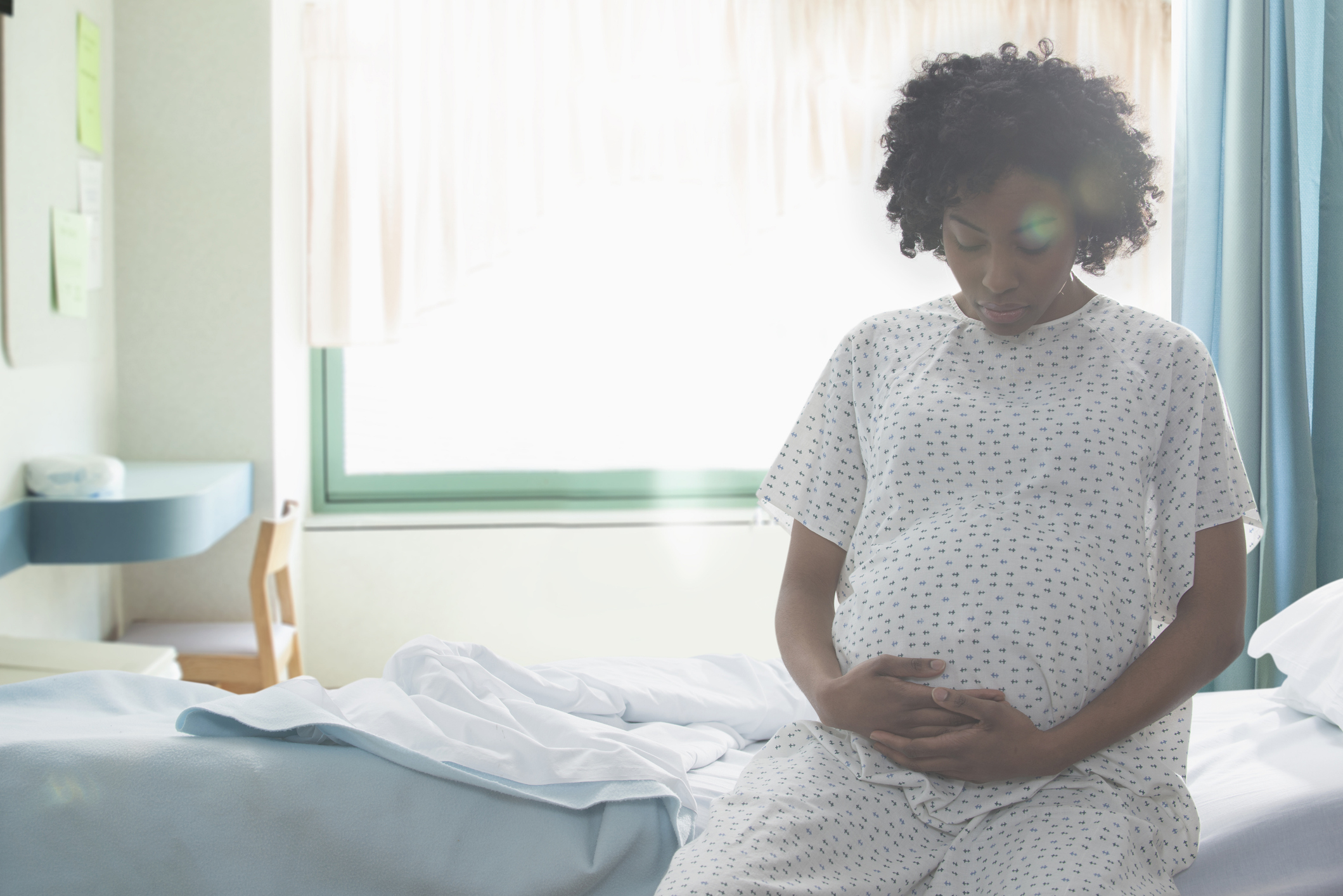 Pregnant person sitting on hospital bed, resting hand on belly, contemplative expression, hospital gown worn