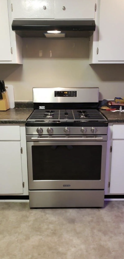 A stainless steel gas range oven is centered between white kitchen cabinets with a range hood above