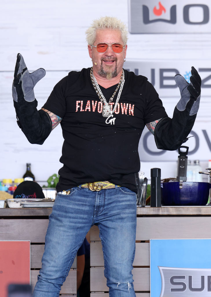 Guy Fieri poses excitedly on stage with his hands up wearing a Flavortown shirt and jewelry