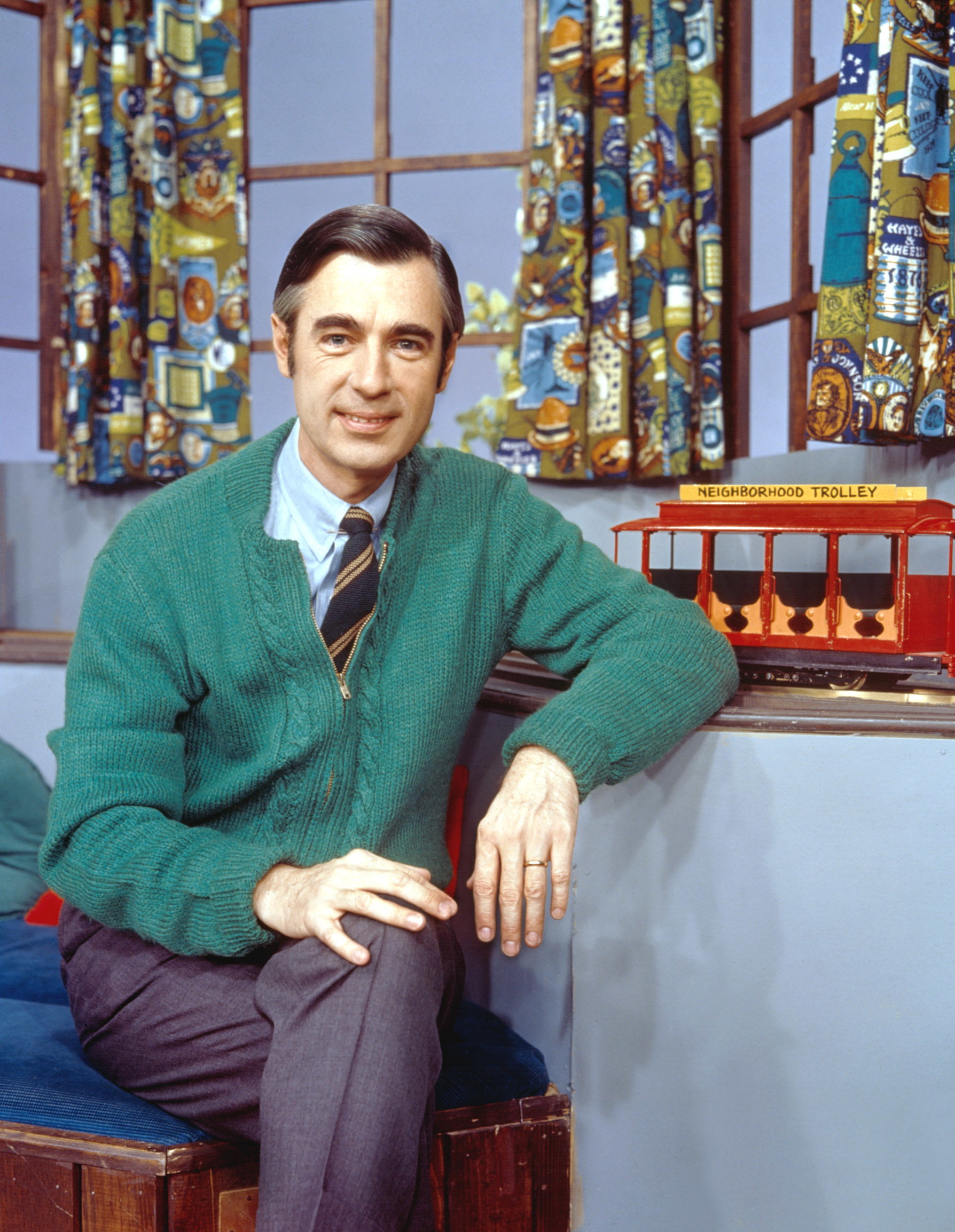 Fred Rogers in a green cardigan sits with a trolley model from his show