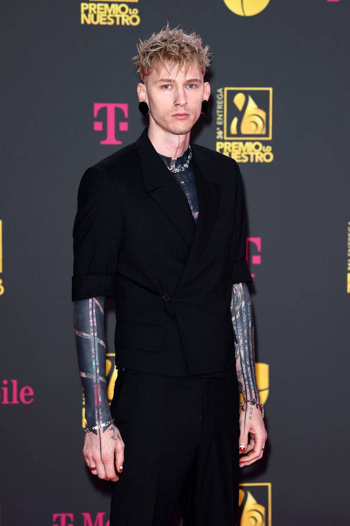 Person stands on the event backdrop, wearing a structured black jacket with tattooed arms visible