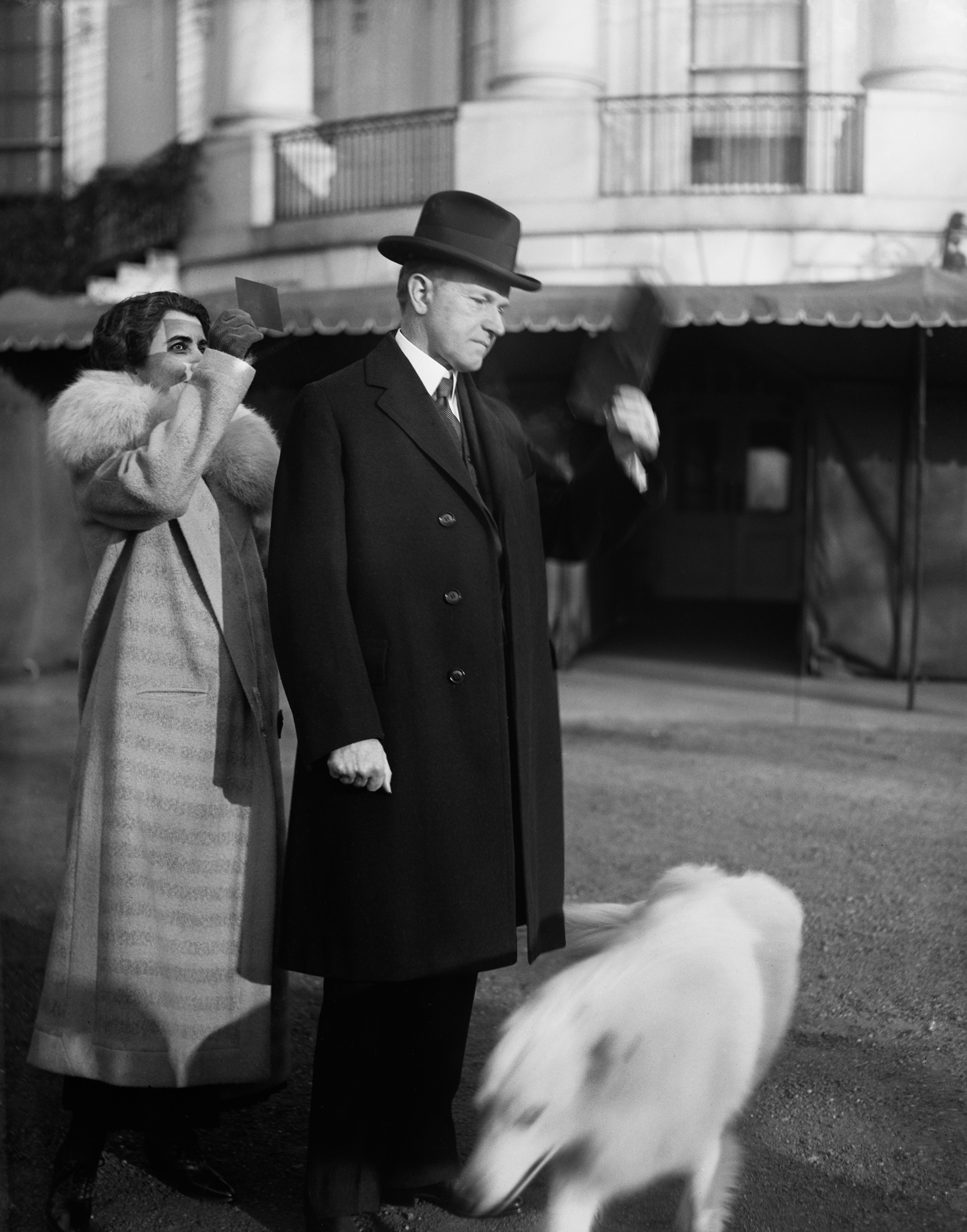 Woman in fur coat and man in suit and hat walking, blurred motion, dog in foreground