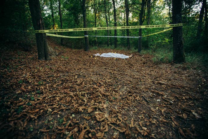 A covered body lies at a crime scene in the woods with yellow police tape surrounding the area
