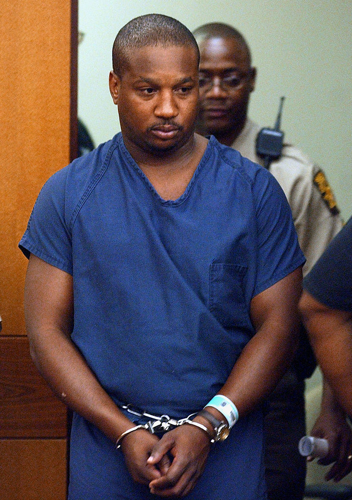 A handcuffed man in a blue jumpsuit escorted by an officer in court