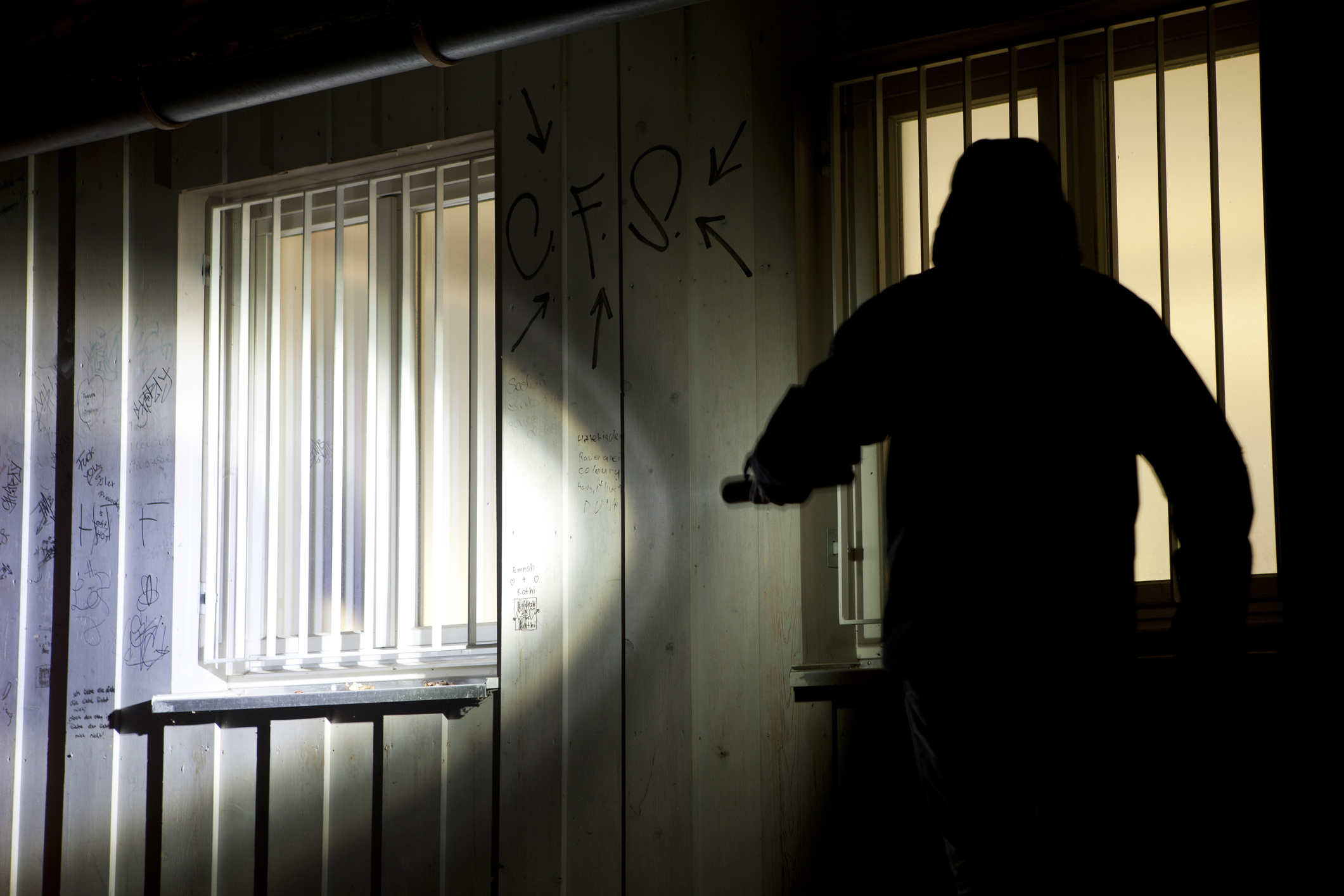 Silhouetted figure pointing a flashlight at a graffiti-covered wall with barred windows