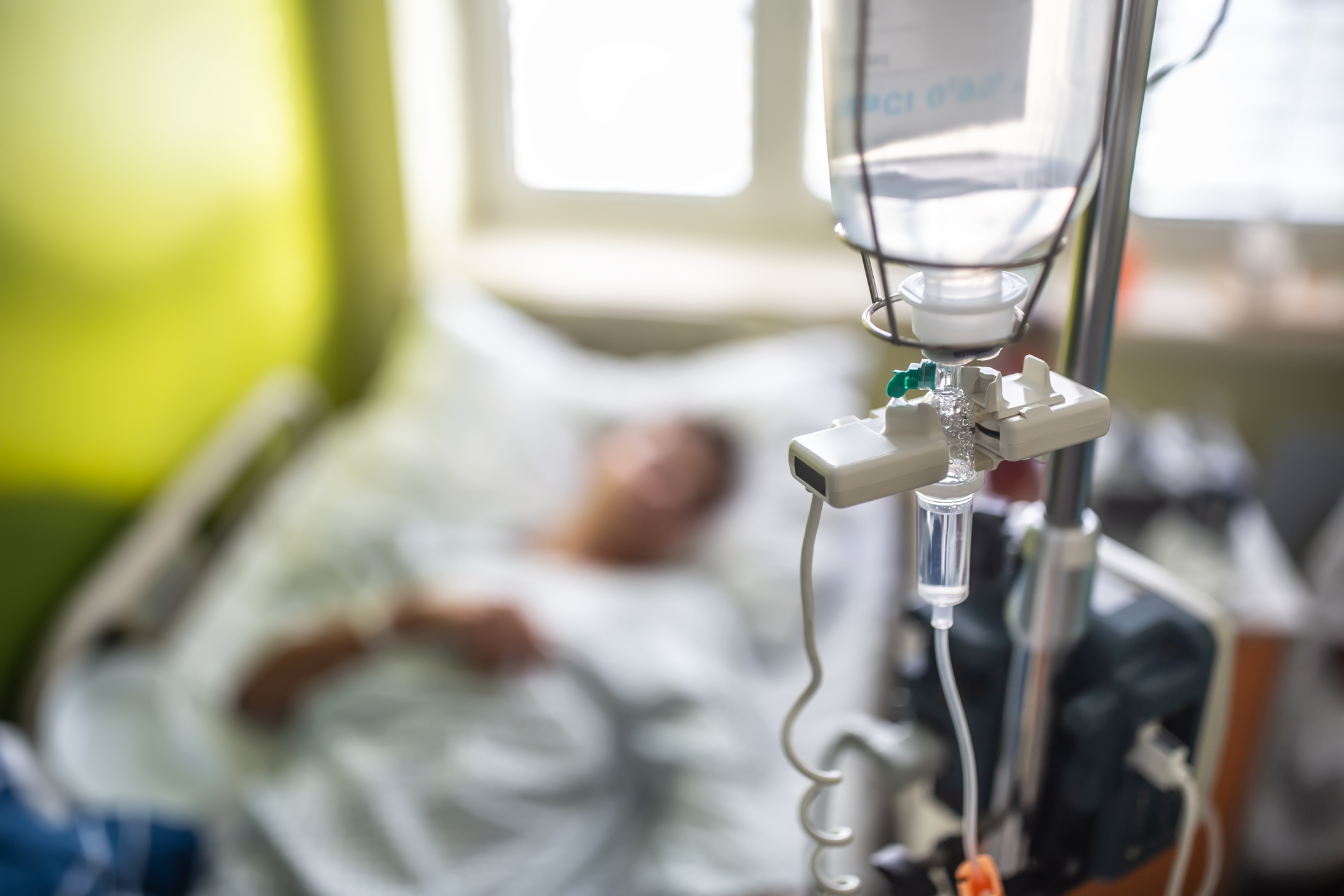 A hospital room with an IV drip in the foreground and a patient resting in the background