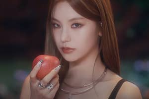 Woman holding an apple, adorned with rings and a layered necklace, gazing at the camera