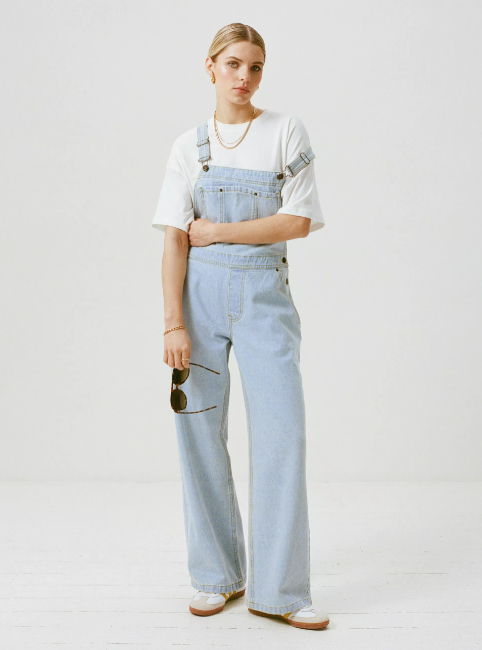 model in denim overalls over a white tee, accessorized with hoop earrings and sunglasses, standing casually