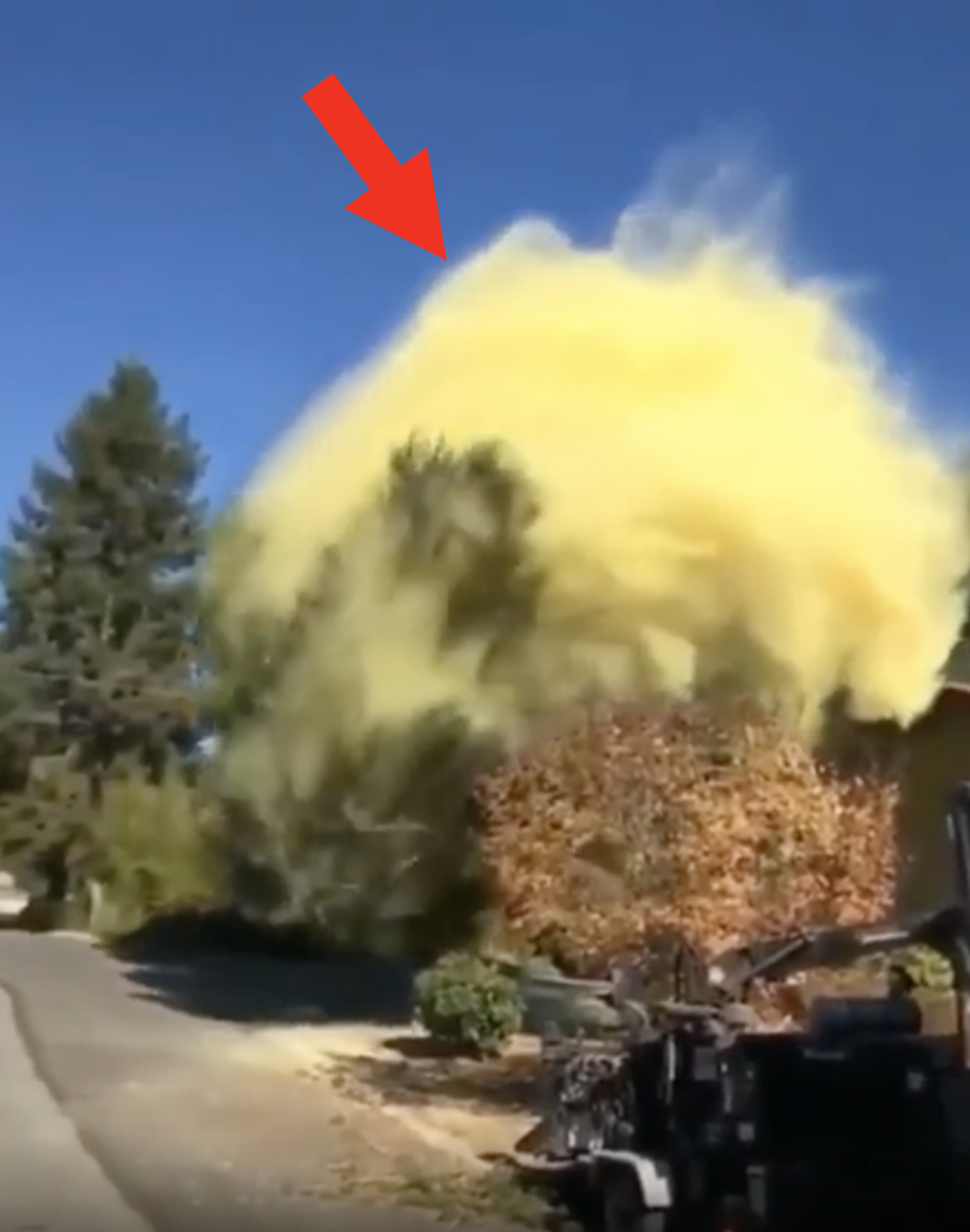 A large yellow smoke cloud billows from a device on a residential street, with trees and a clear sky in the background