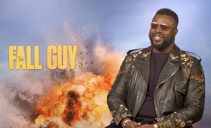 Man sitting, wearing floral jacket and sunglasses, with &quot;FALL GUY&quot; text and explosion graphic in background
