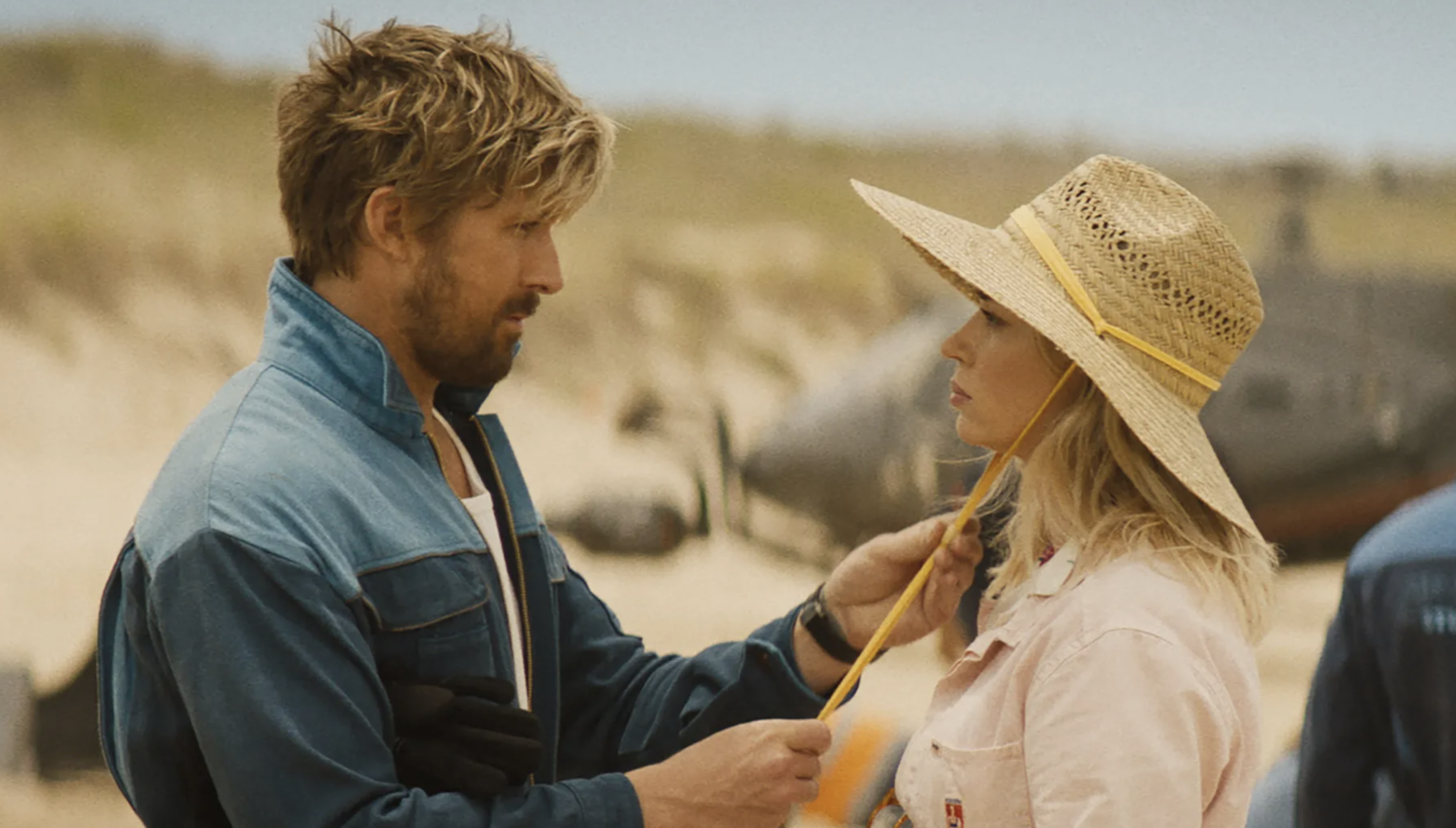 Man in denim jacket talking to woman in straw hat and pastel shirt on beach setting