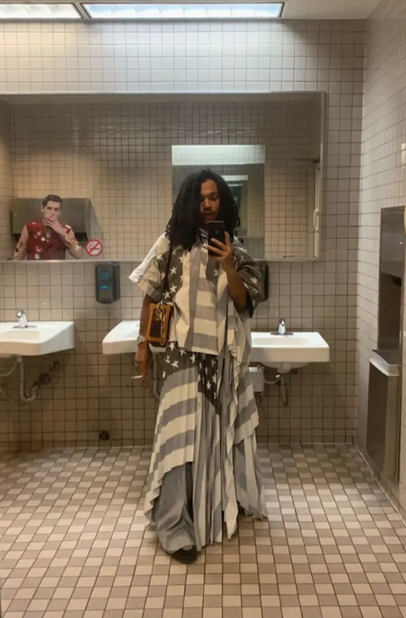 Luka in a patterned outfit takes a mirror selfie in a restroom with another person in the background