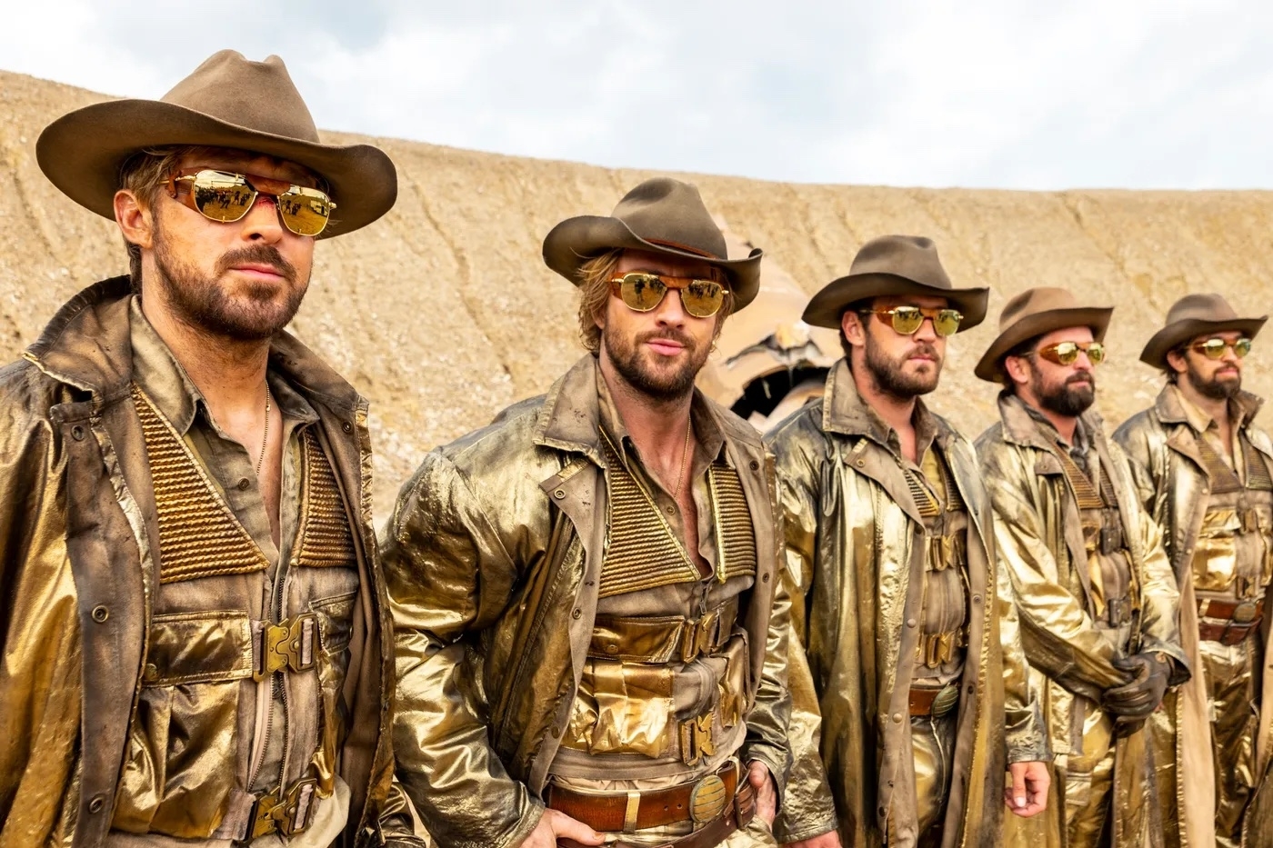 Five actors in cowboy outfits with shiny gold accents, wearing hats and sunglasses, posing with a desert backdrop