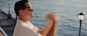 A man in a white shirt and sunglasses claps his hands outdoors near water