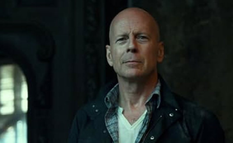 Bruce Willis in character wearing a casual jacket and shirt. He&#x27;s indoors, looking serious