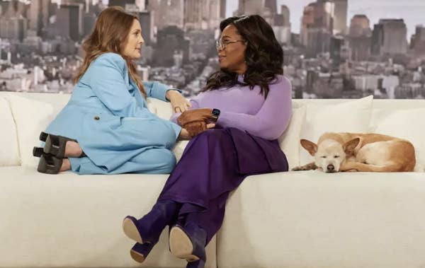 Two women in conversation on a couch, one with a dog beside her, on a talk show set
