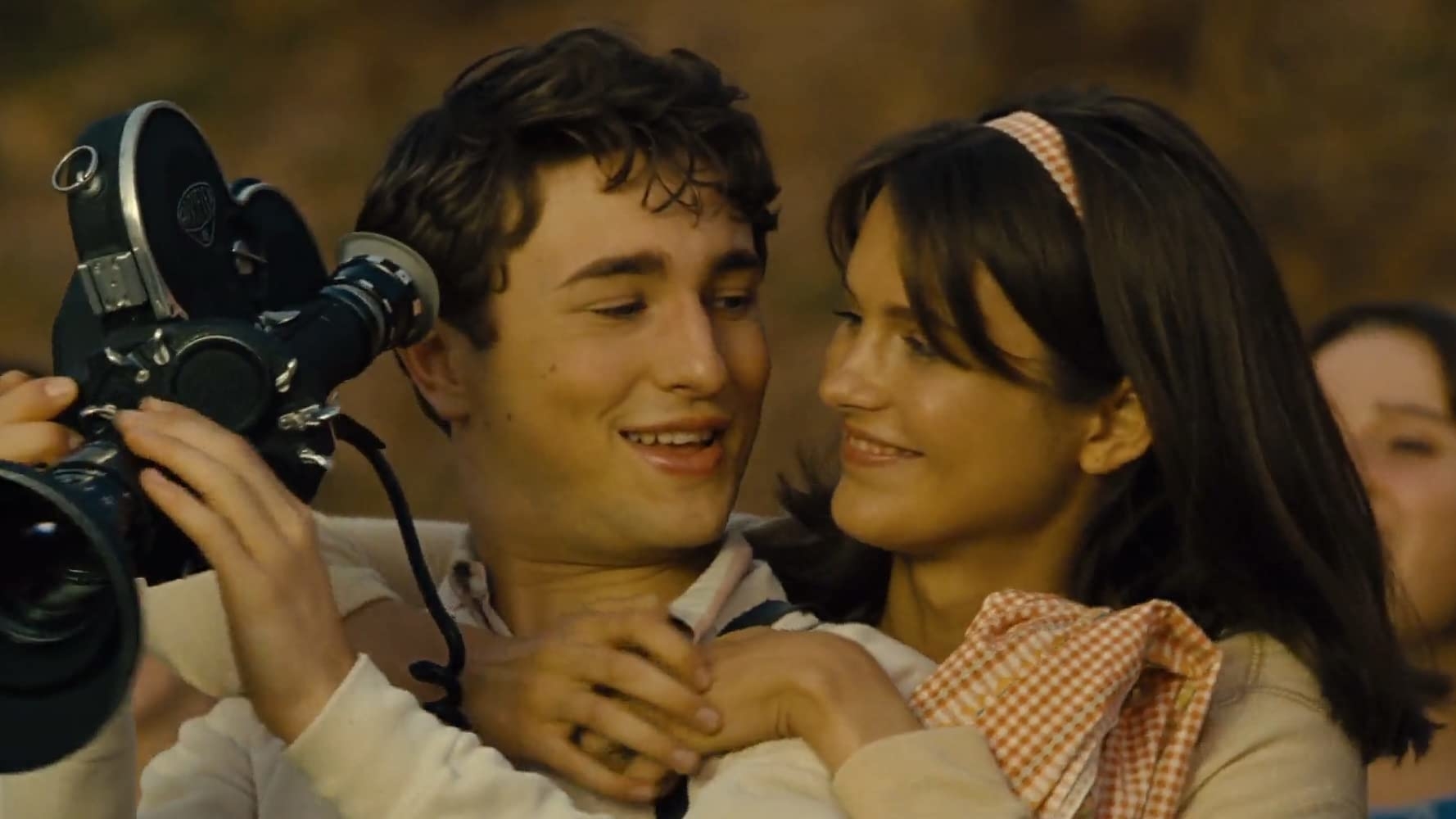 Two characters share a joyful embrace while holding a film camera, suggesting a scene from a movie or series