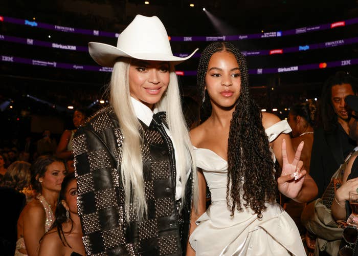 Two women at an event, one wearing a white hat and black embellished jacket, the other in an off-shoulder satin gown