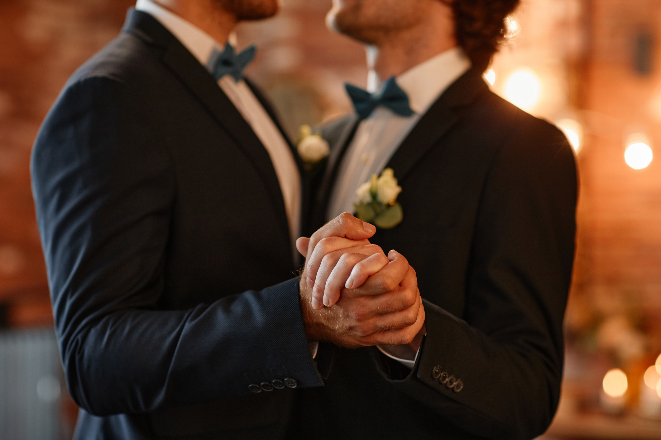 Two individuals in suits with bowties holding hands, likely in a wedding ceremony