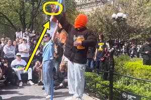Man in orange face mask holding up an object in a crowded park with arrow pointed to it