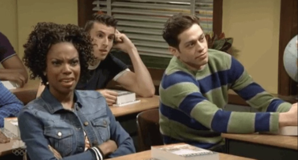 Scene from a TV show with three characters seated at a desk, displaying various expressions of skepticism and curiosity