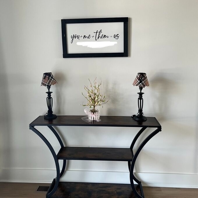 Wall-mounted framed art with the phrase &quot;you + me = them us&quot; above a console table with two lamps and a vase