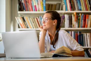 Woman at desk with laptop and book, looking thoughtful