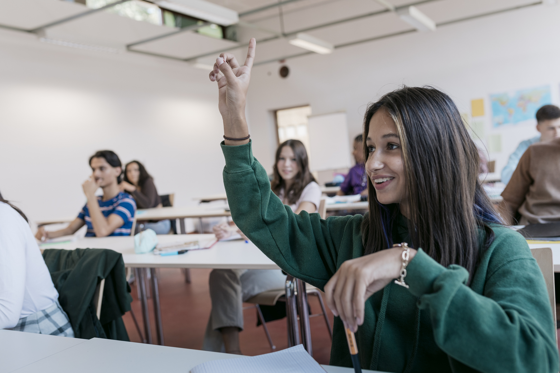 Student raises hand in classroom with peers, engaging with the teacher off-camera