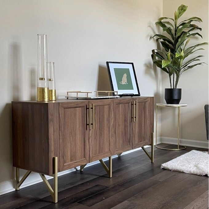 Wooden credenza with brass legs and handles, decorative items on top, next to a plant on a stand