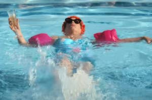 Person in a pool with floaties, holding a glass and wearing sunglasses