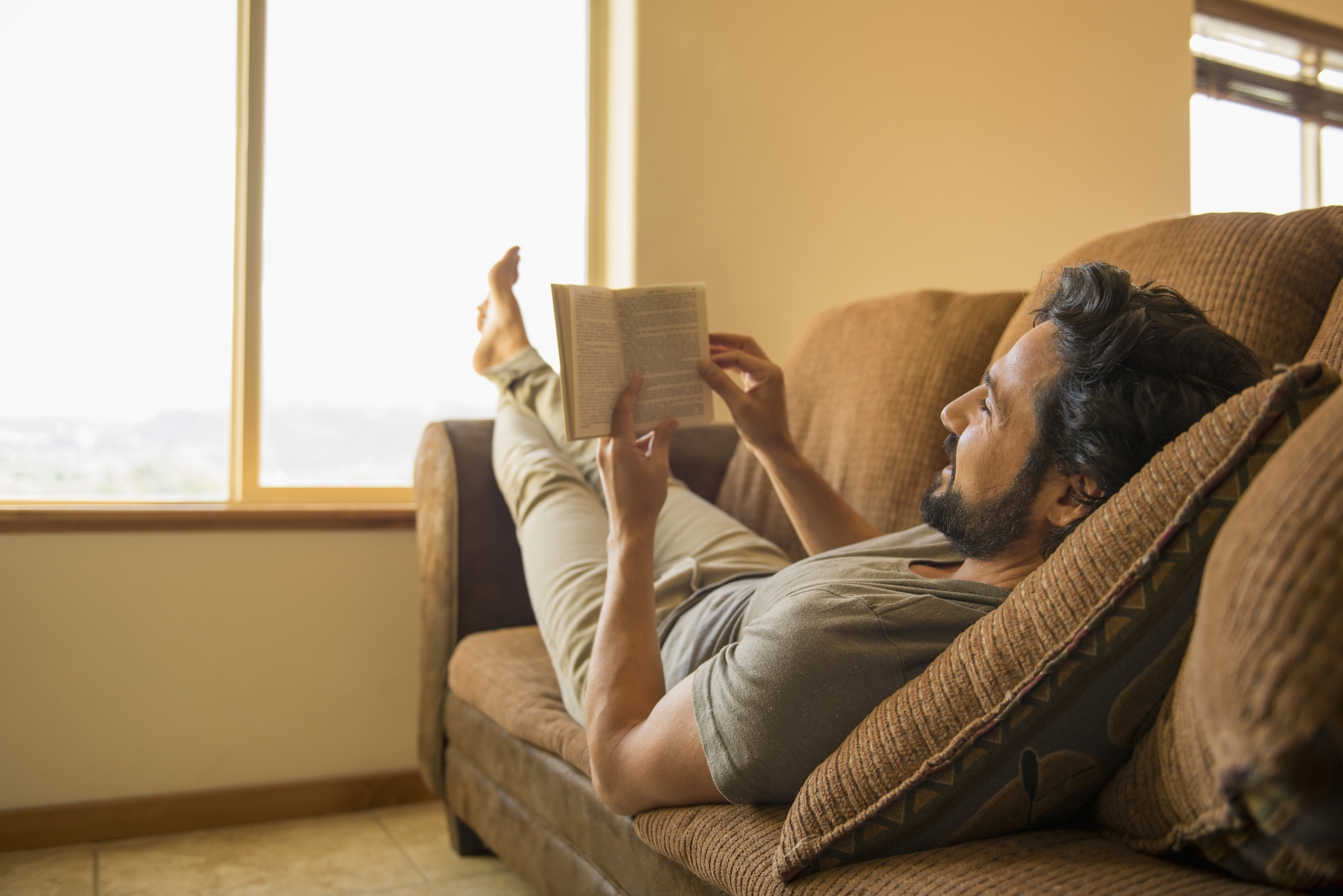 Person lounging on a sofa reading a book by a window, relaxed setting