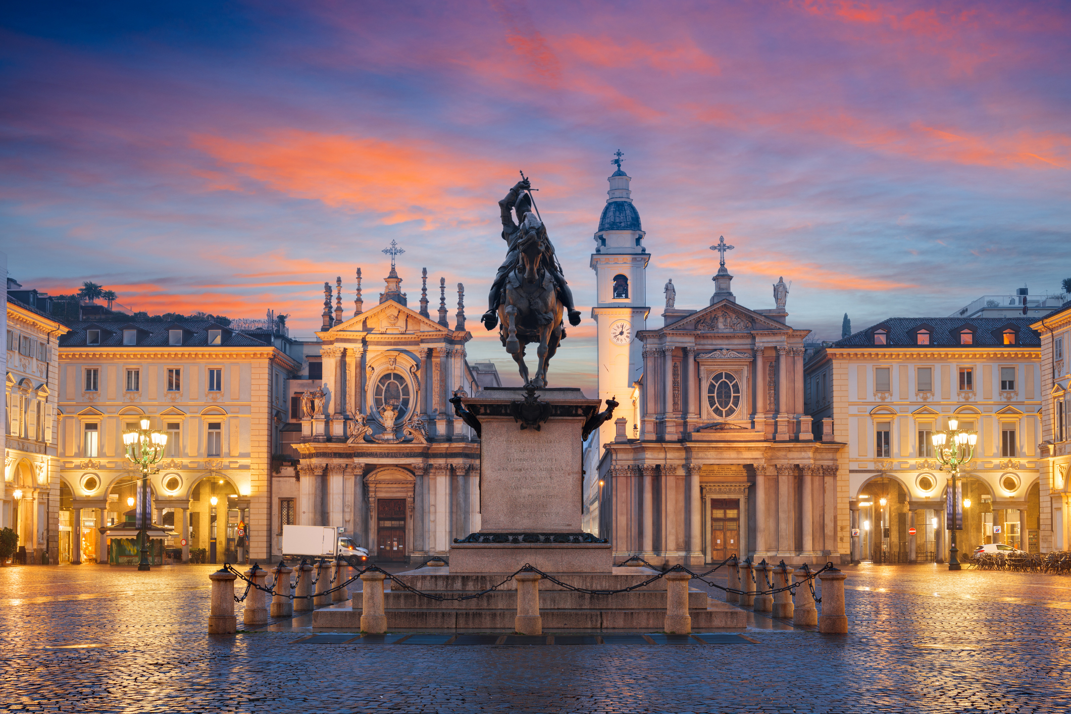 Historical square with a statue and two churches under a sunset sky