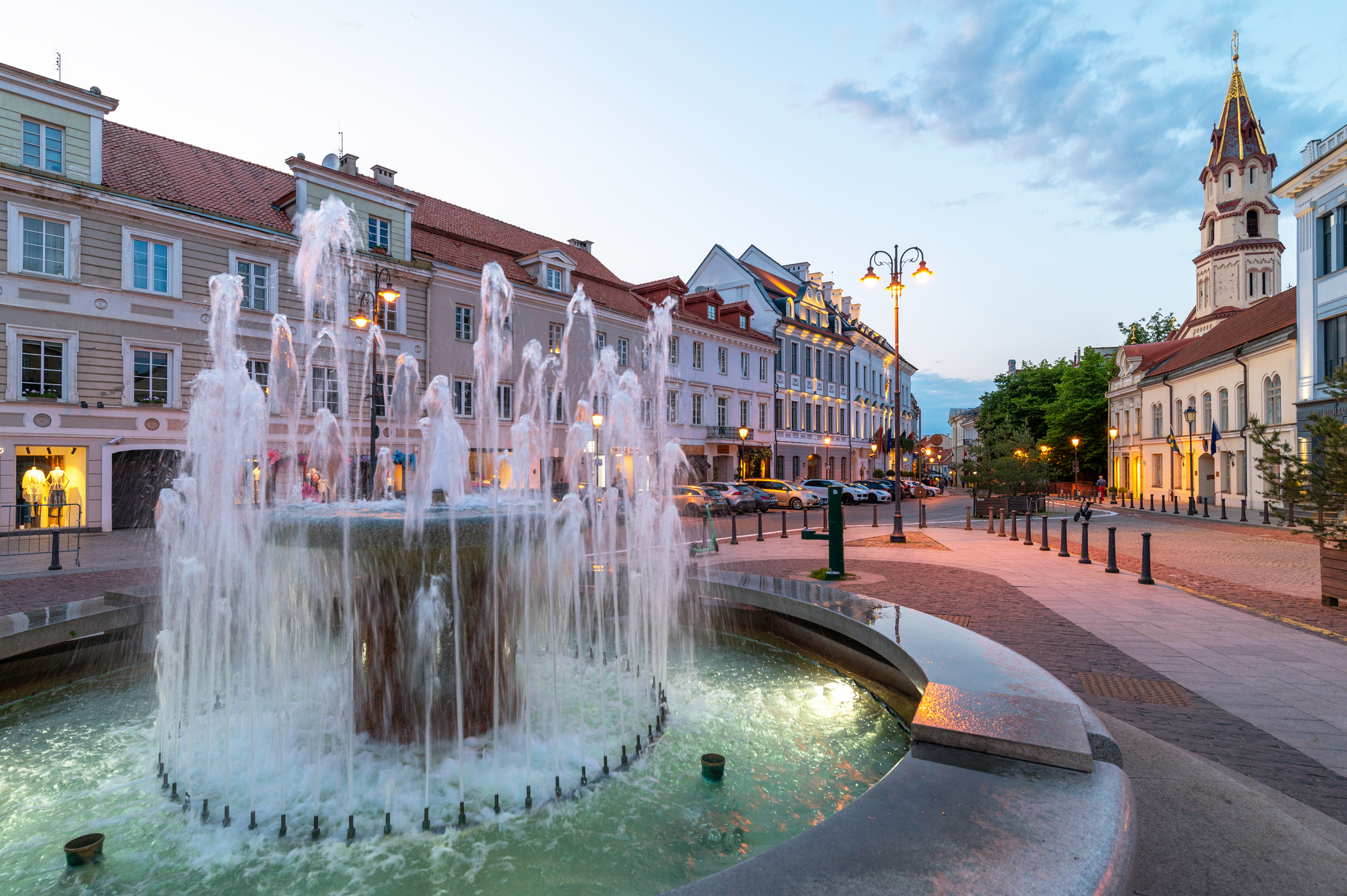 Fountain in a city square during twilight with surrounding buildings and a tower in the background