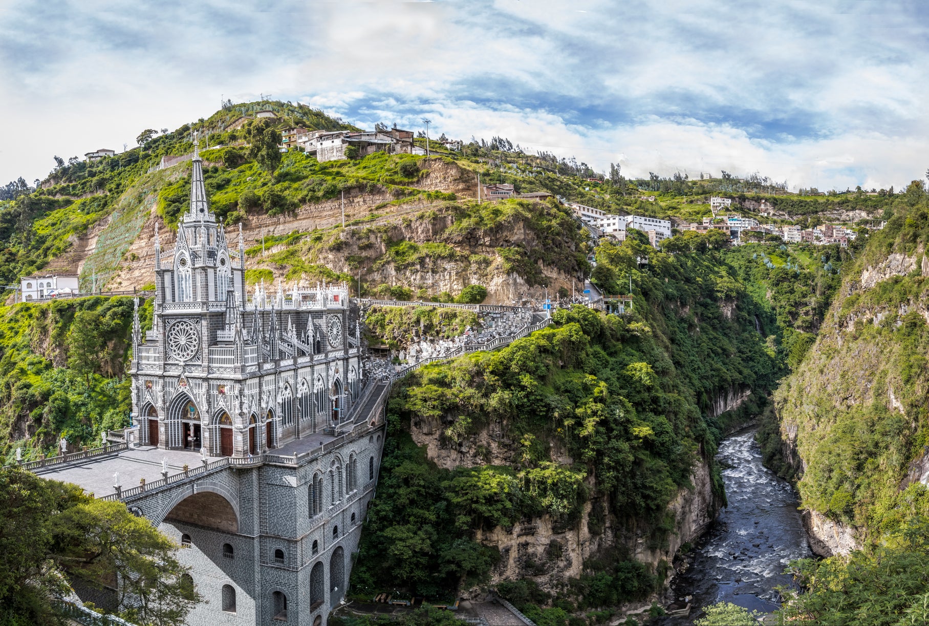 Gothic-style church perched on cliff edge beside a river, with surrounding greenery and buildings on the hilltop