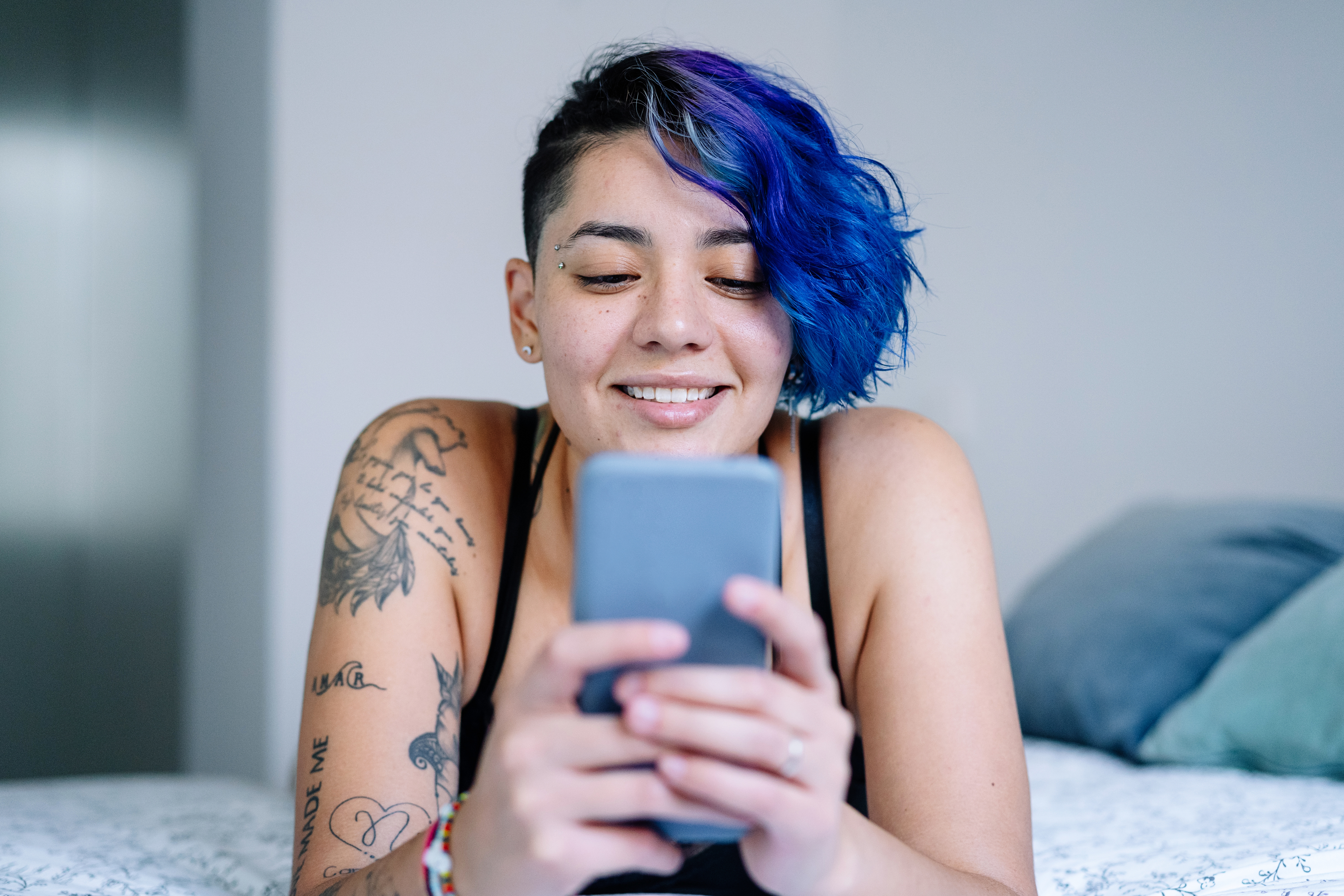 Person with blue hair smiling, holding a smartphone, tattoos visible on arms