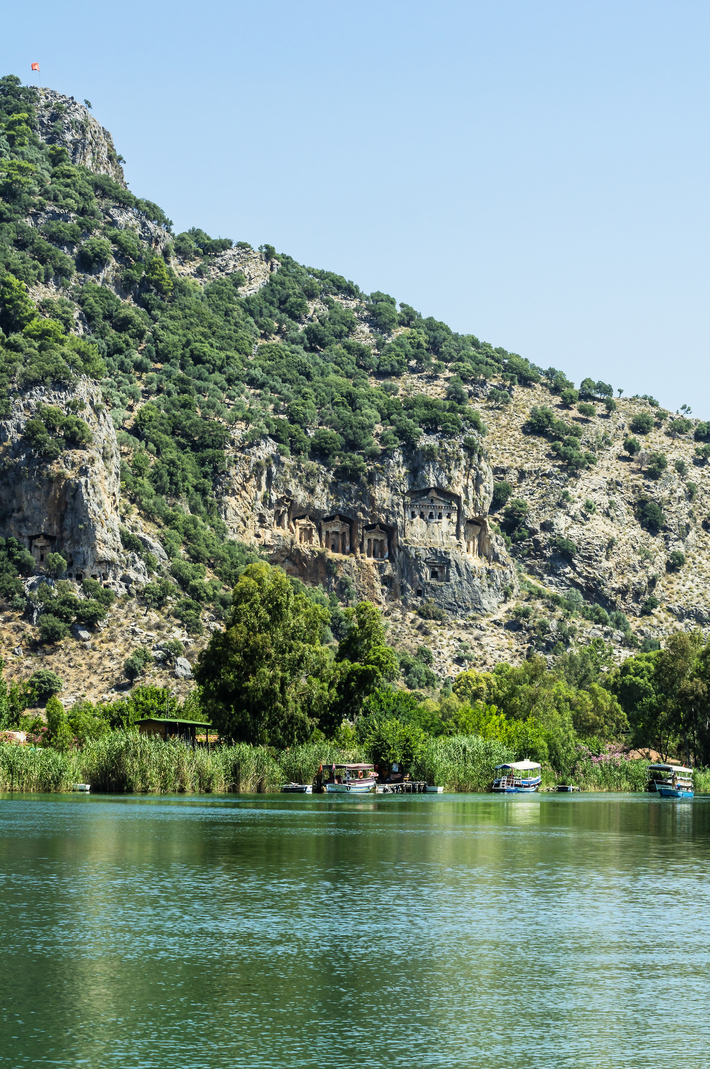 Scenic view of a calm river with boats, bordered by lush greenery and rocky cliffs with ancient carvings