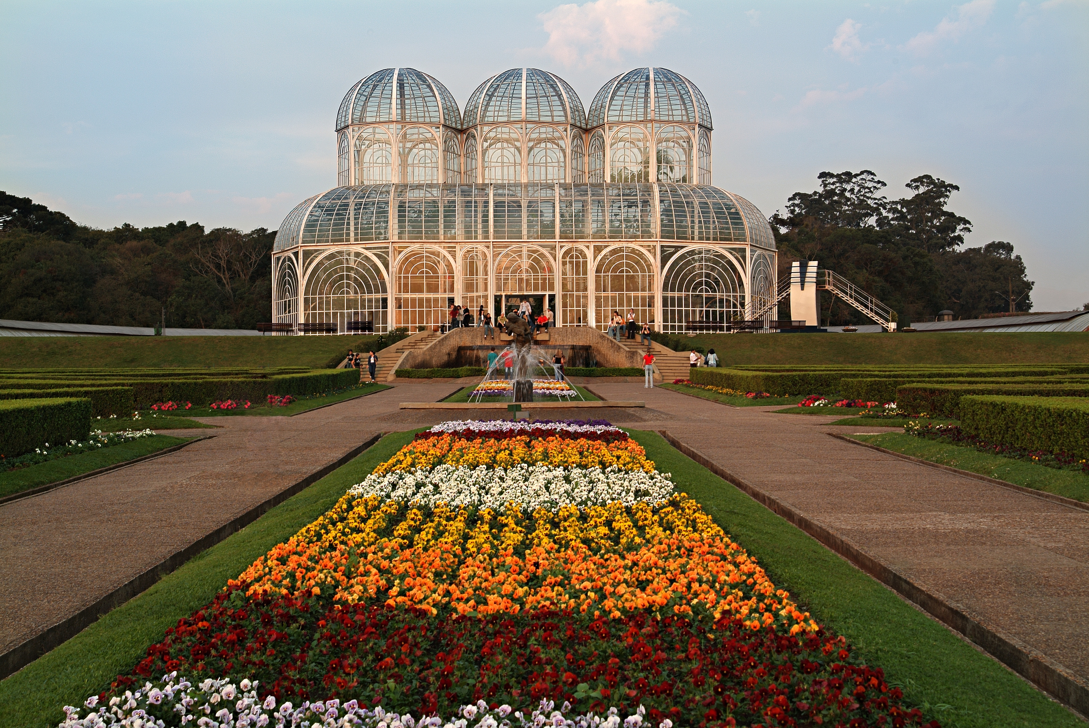 Glass greenhouse with a geometric design, flanked by symmetrical flower beds and a fountain in front