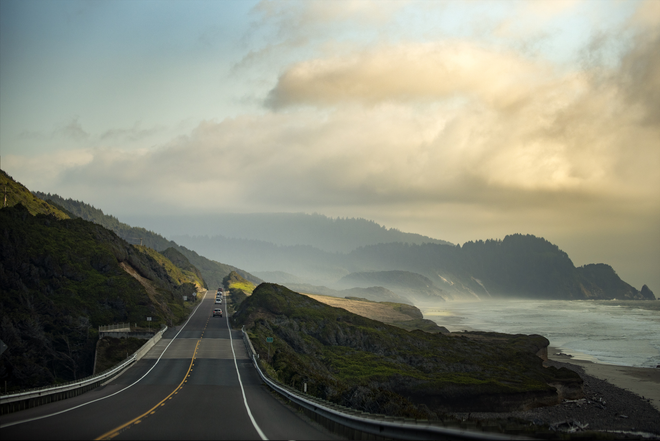 Coastal highway with vehicles, ocean on one side, hills and cloudy skies above