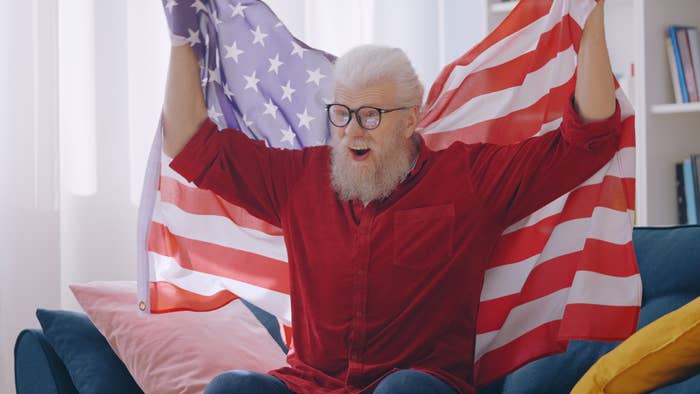 Elderly man with beard holding up an American flag indoors, expressing enthusiasm