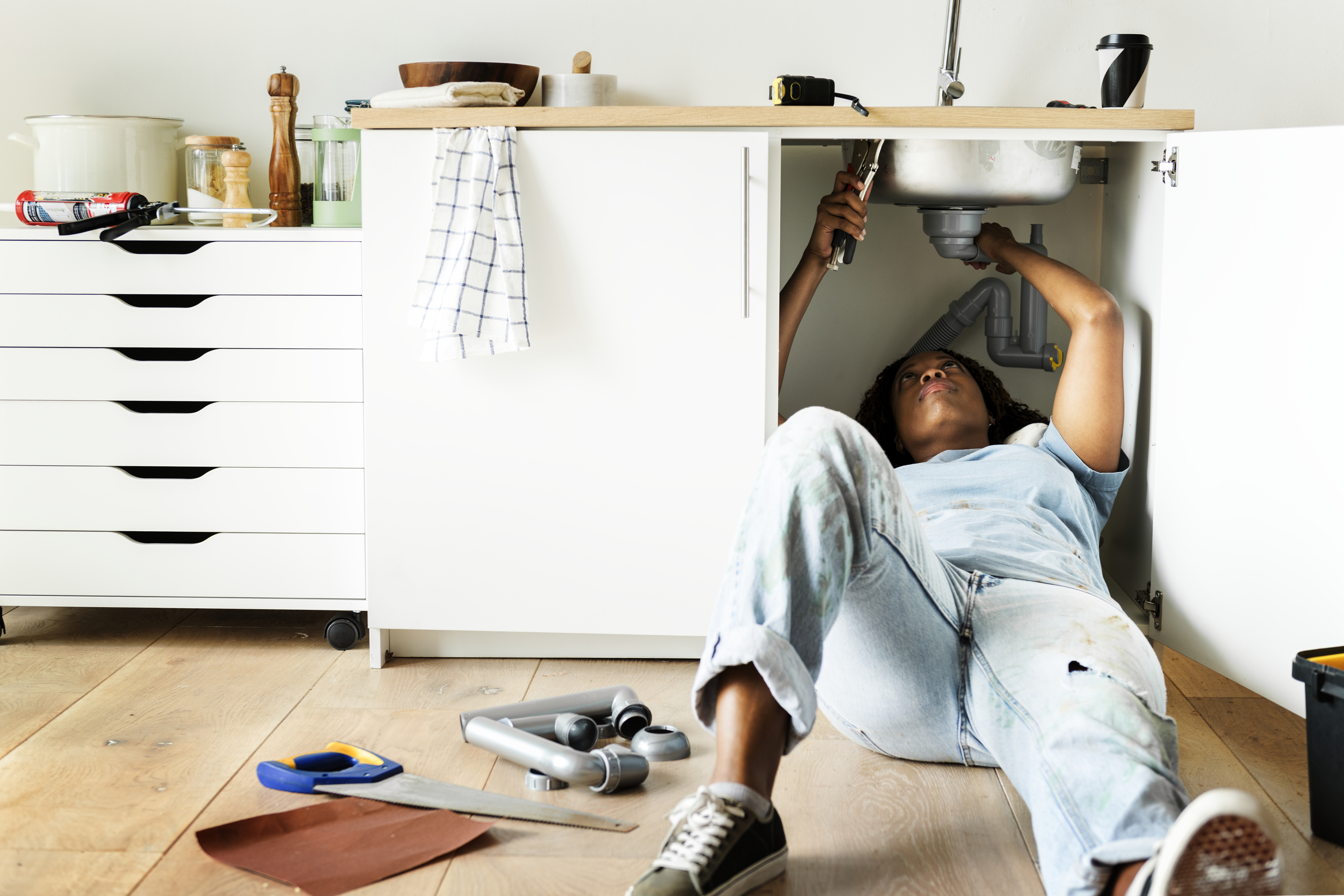 Person lying on floor using a wrench under a sink, with tools scattered around