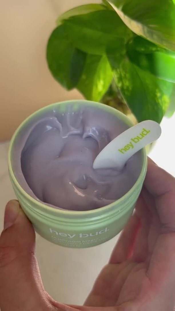 Hands holding a jar of Hey Bud skincare cream with a spatula resting on top, green plant in background