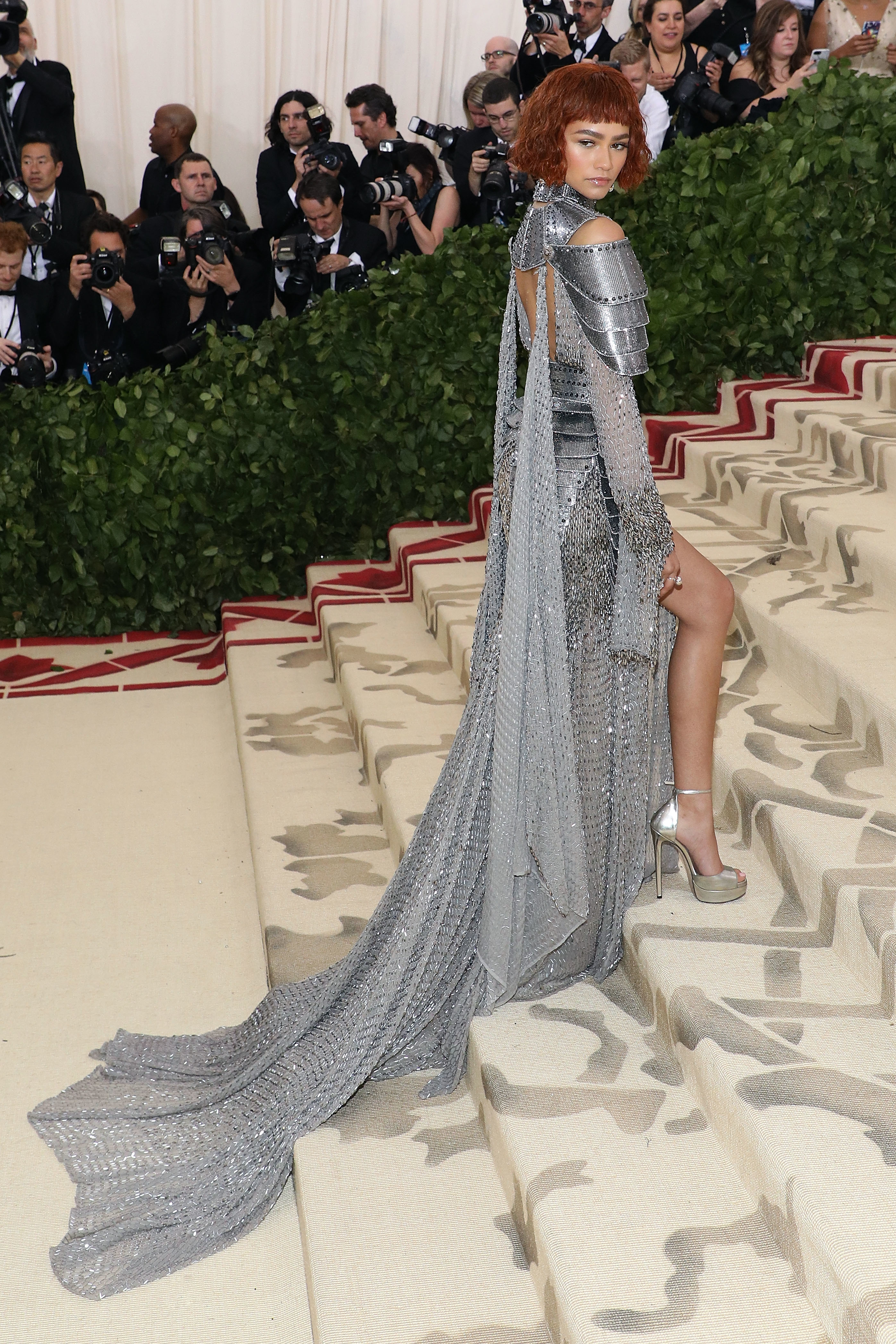 Zendaya in a metallic gown with a long train and arm armor posing on stairs at the Met Gala
