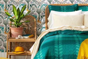Rattan bedside table with plant and book beside a bed with textured teal and white bedding in a room with patterned wallpaper
