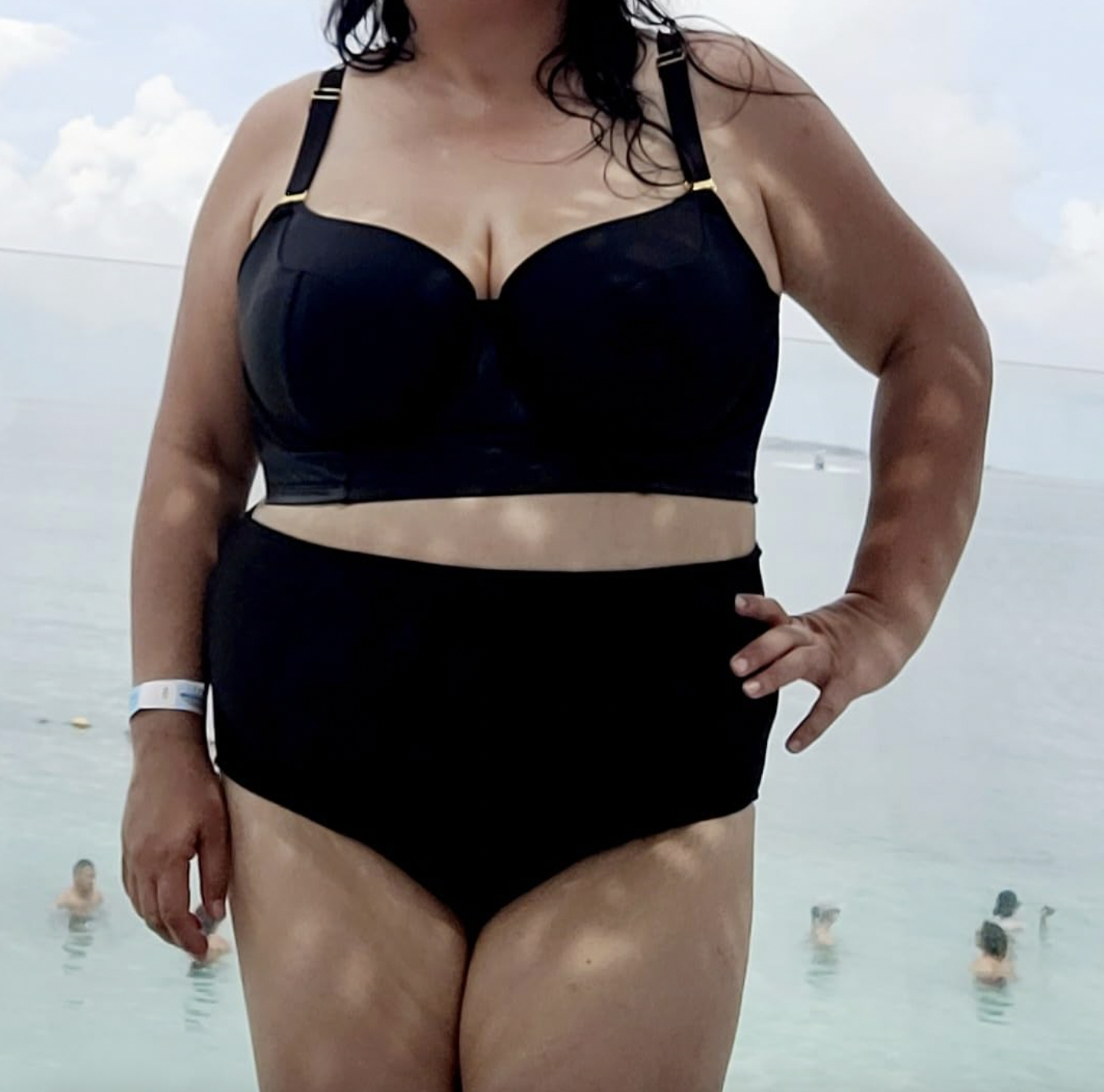 reviewer in black two-piece swimsuit at beach, hands on hips, others swimming in background