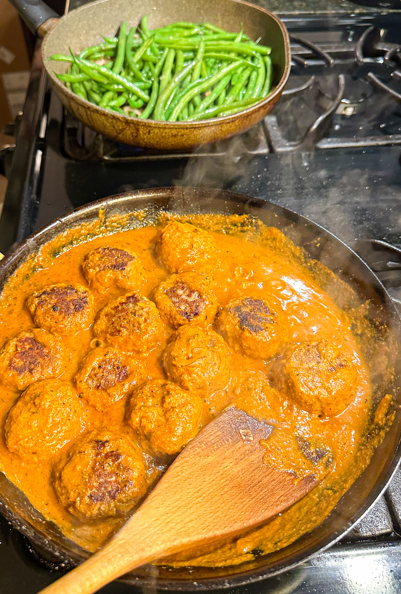 Meatballs simmering in a sauce with a pan of green beans in the background on a stove