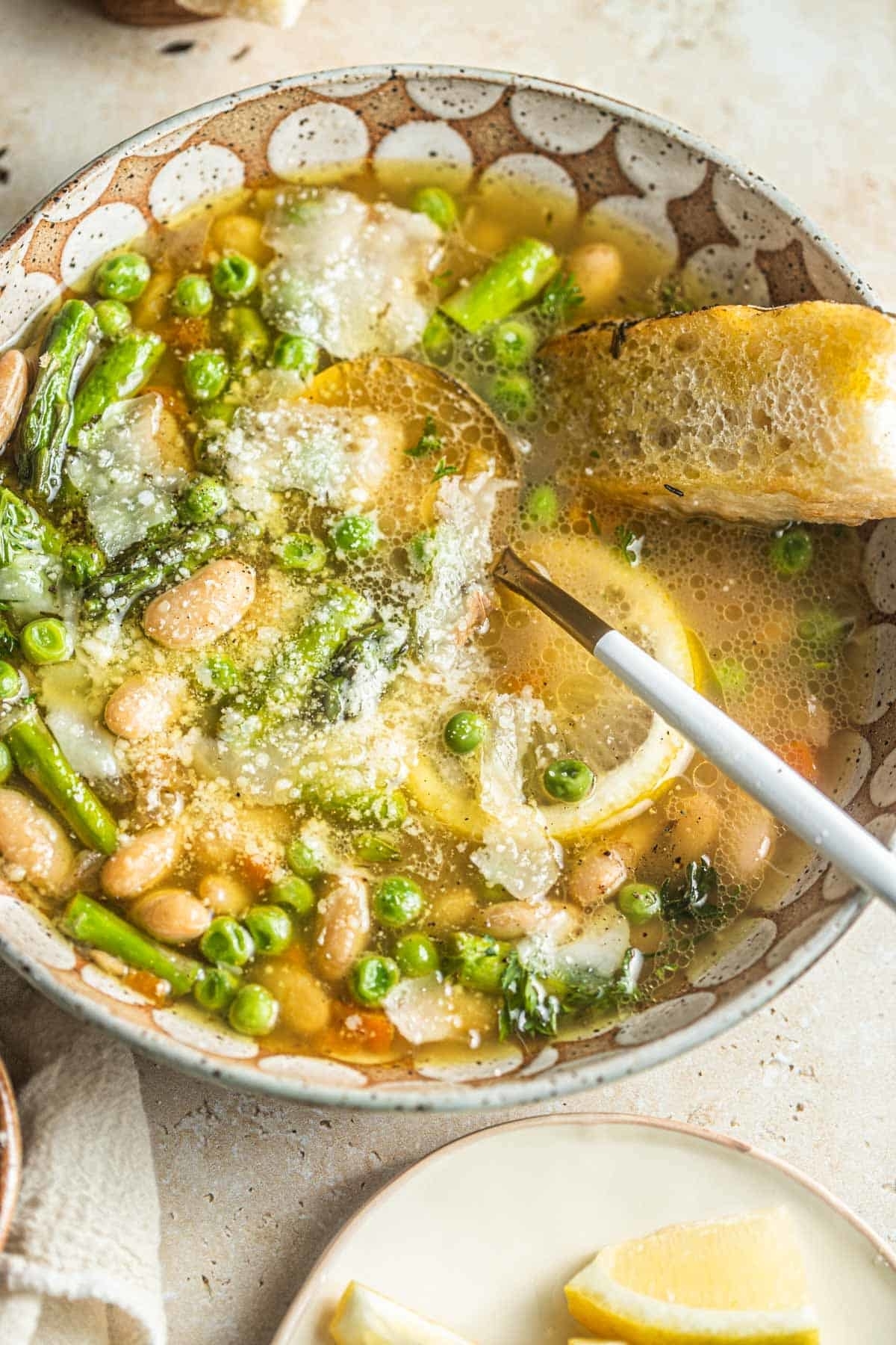 Bowl of soup with beans, peas, asparagus, lemon slices, and topped with grated cheese